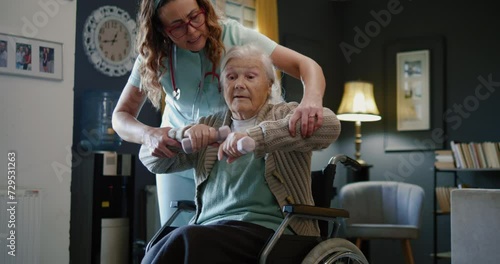 Female nurse physiotherapy worker help elderly woman with disability to exercise with dumbells, caregiver assist a senior woman in wheelchair to do physical therapy and exercise at home photo