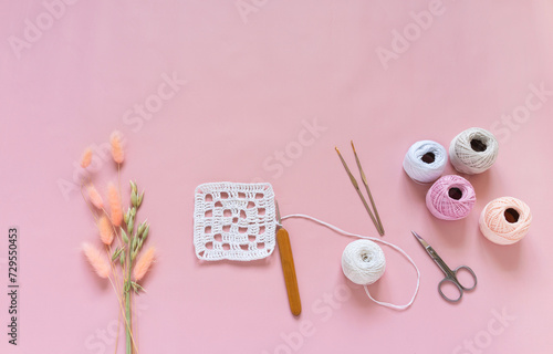 Top view of crocheting process of white square lace doily on pink pastel background. Skeins of yarn for knitting, crochet hooks and bouquet of ears of oats and lagurus nearby. Copy space, flat lay