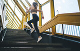 Dynamic young Caucasian woman sprinting up the stairs in an architecturally striking yellow overpass, promoting endurance and agility in a summer fitness regimen