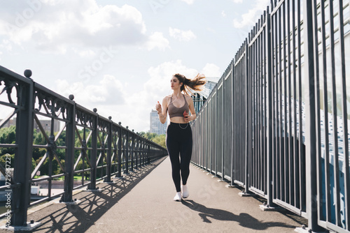 Confident young adult Caucasian woman jogging on a city bridge, wearing sportswear with earphones, enjoying a sunny day run, cityscape in the background © BullRun