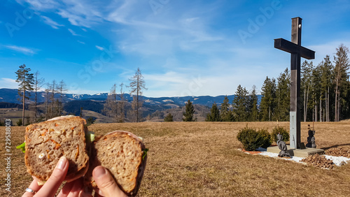 Eating sandwiches at summit cross on alpine meadow in West Styrian highland, Voitsberg, Styria, Austria. Hiking in remote landscape of soft hills in spring. Scenic view of mountains of Lavanttal Alps photo