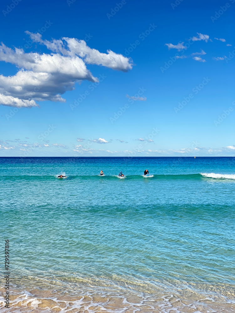 Surfers practicing catching a wave in the turquoise-blue ocean. Surf lesson. A day at the beach on a sunny day with blue sky and clouds. Surfers practicing catching a wave in the turquoise-blue ocean. Surf lesson. A day at the beach on a sunny day with blue sky and clouds.