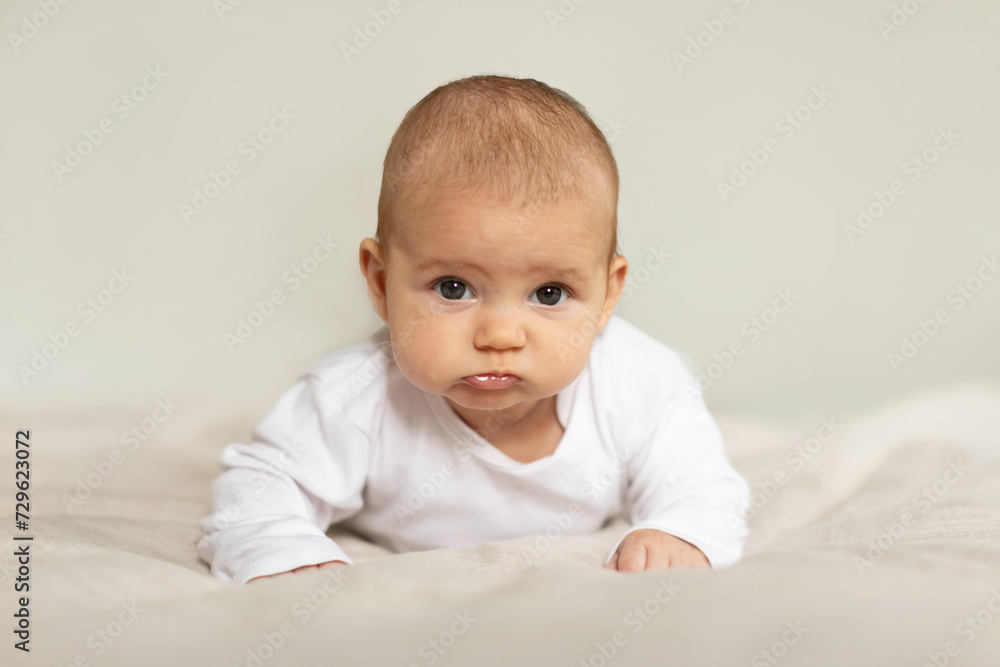 Adorable baby girl in white bodysuit lying on bed, child relaxing in bedroom, infant kid during tummy time, free space