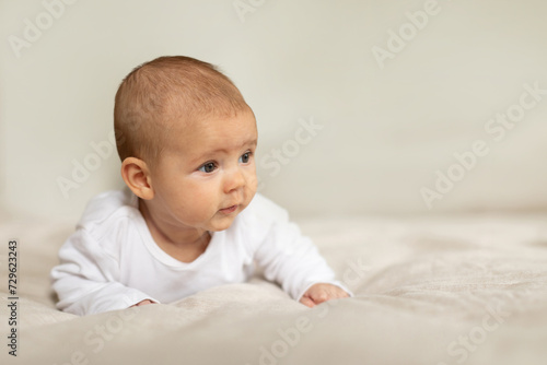 Baby girl wearing white clothes lying on bed in light nursery room, infant child girl relaxing in bedroom, free space