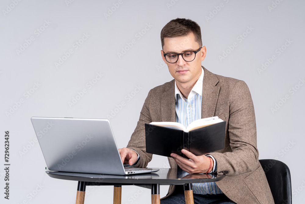 Young man sitting at the table and working on laptop and book on gray background Young man sitting at the table and working on laptop and book on gray background