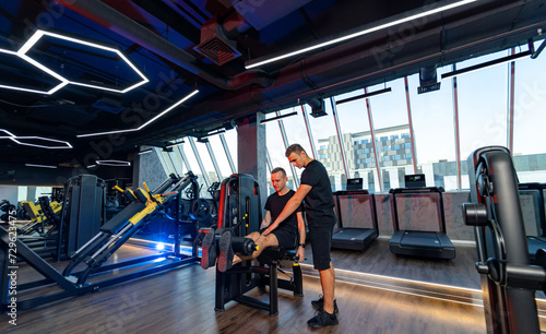 Personal trainer helping young man to do leg exercises with leg press machine in the modern gym interior