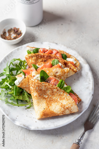 Crepes or thin pancakes with smoked salmon, soft cheese and spinach on a plate on gray textured background. Pancake day, Maslenitsa