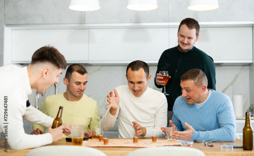Carefree emotional male friends playing fun drinking board game with shot glasses replacing pieces during bachelor party at home..