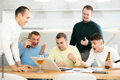 Group of male friends gathering at home enthusiastically celebrating good news, seen on laptop screen, with lively gestures and drinks..