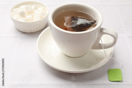 Tea bag in cup with hot drink and bowl with sugar on white tiled table, closeup
