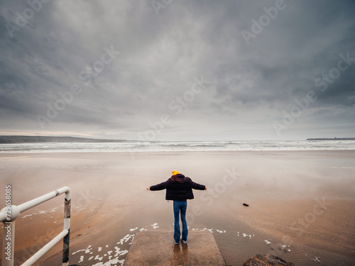 Teenager girl in black jacket standing on a footpath to a vast sandy beach and ocean, dramatic sky in the background. Trip to nature concept. Lahinch, county Clare, Ireland. Travel and tourism theme.