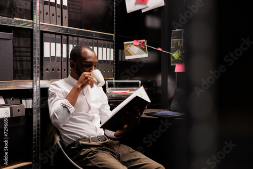 Detective drinking coffee and studying forensic report, searching insight to solve crime. African american investigator reading crime case file and analyzing evidence in office at night