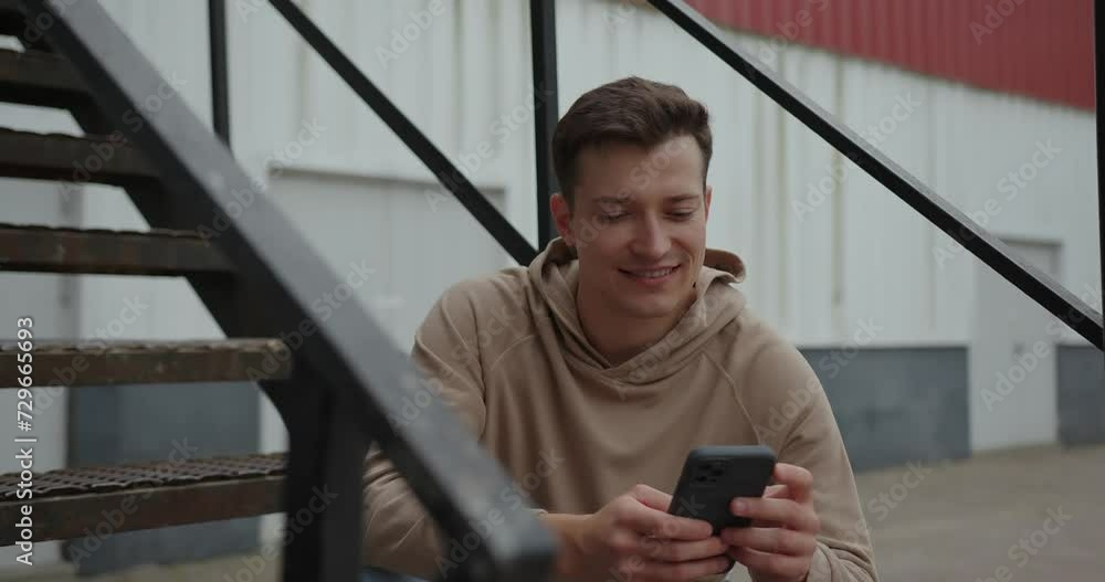 A young man is sitting on the metal stairs on the street, holding the phone in his hands. A young man is sitting on the metal stairs on the street, holding the phone in his hands.