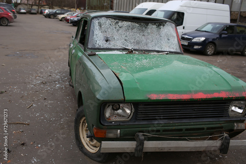 An abandoned green car with a shattered windshield parked on an urban lot, showcasing signs of neglect and vandalism.
