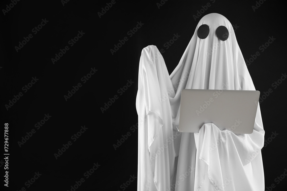 Creepy ghost. Person in white sheet with laptop on black background, space for text Creepy ghost. Person in white sheet with laptop on black background, space for text