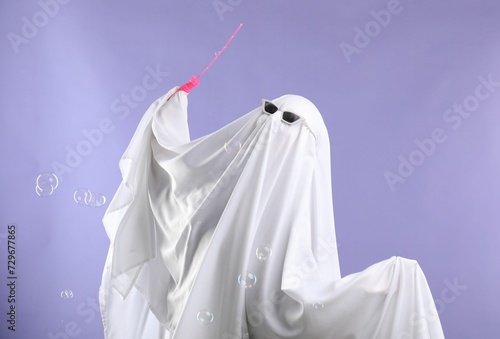 Funny ghost. Person in white sheet, sunglasses and soap bubbles on violet background