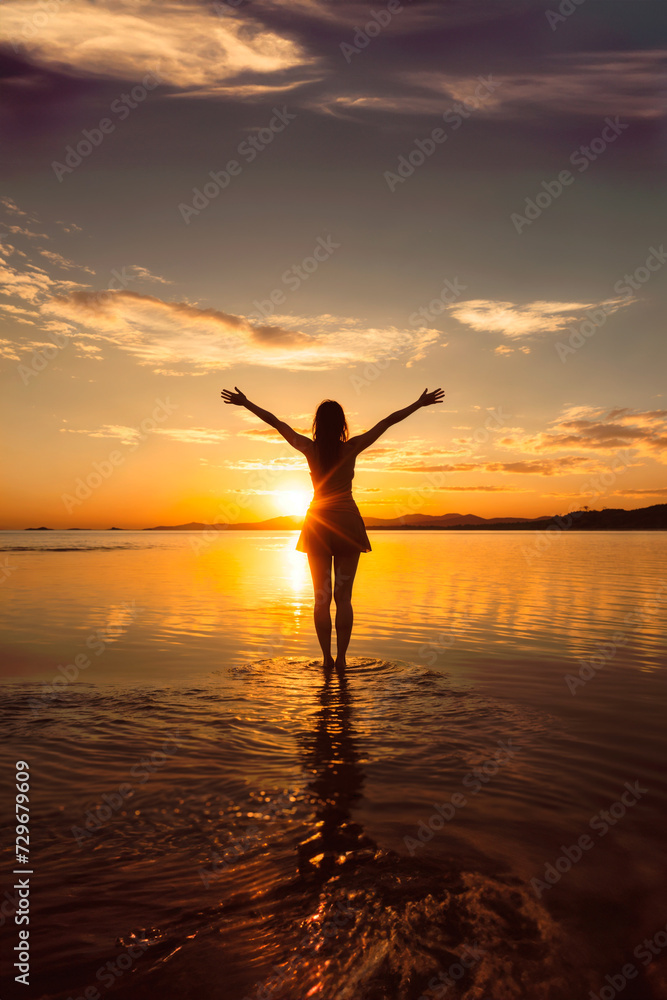 Saluting the sun. Backlight of a woman on the beach facing the sea, with her arms open, contemplating the sunset. AI Generated.