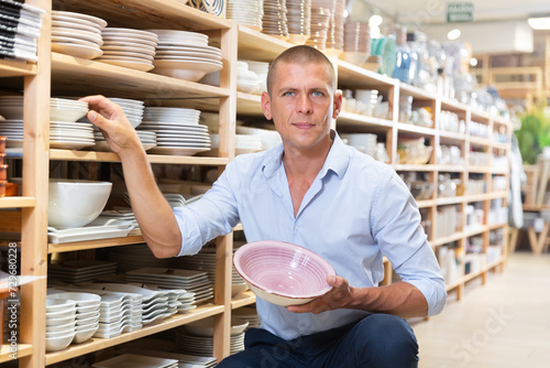 Young cheerful man customer chooses plates in tableware store