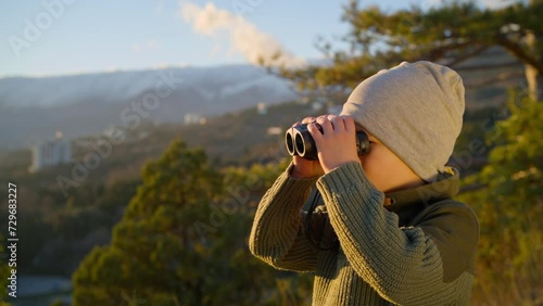 Boy looking through binoculars around while in nature, in the mountains in the rays of the setting sun on a spring evening.