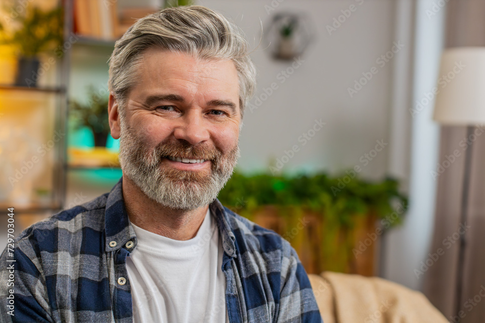 Happy Caucasian senior man sitting on sofa looking at camera and smiling at home. Bearded mature guy having attractive appearance enjoying carefree retired life relaxing in living room. Copy-space Happy Caucasian senior man sitting on sofa looking at camera and smiling at home. Bearded mature guy having attractive appearance enjoying carefree retired life relaxing in living room. Copy-space