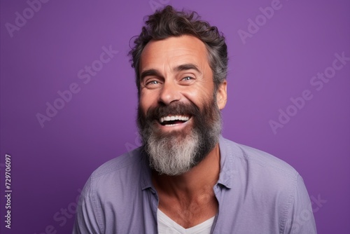 Portrait of happy mature man with beard and mustache looking at camera and laughing while standing against purple background © Iigo