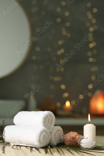 Spa composition. Rolled towels, sea salt and burning candles on table against blurred lights, space for text