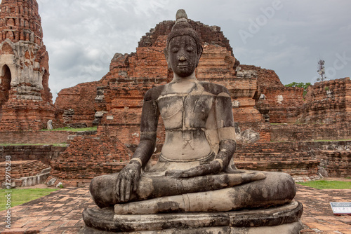 Stone seated buddhist statue and red brick Thai temple pagoda ruin structure and trees of Wat Maha That historical park in Ayutthaya Thailand on a cloudy day