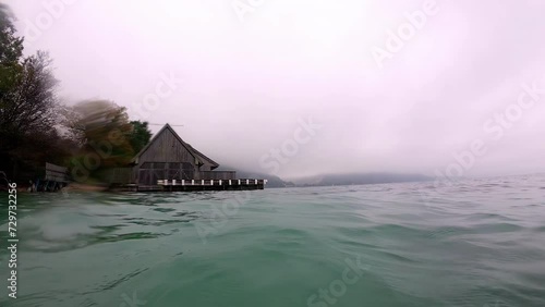Wooden hut on lakeside with mist covered mountains in distance. Waves crashing into camera.  photo