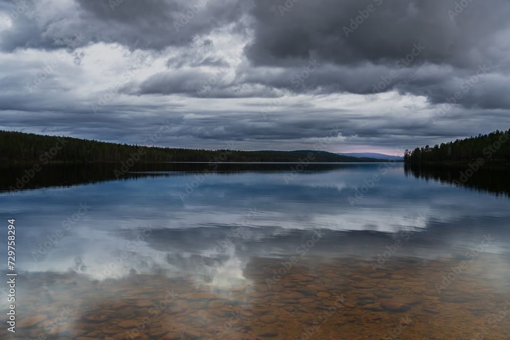 Forest lake at dusk with mountains, pink sky and cloudscape in the background. Clear water with pebbles, reflection of dramatic clouds and trees in the water, in Nordic nature