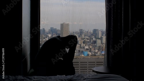 silhouette of adult man anxiously sitting near high rise window, shaking his head in his hands with panoramic view of the city metropolis in background. Static shot