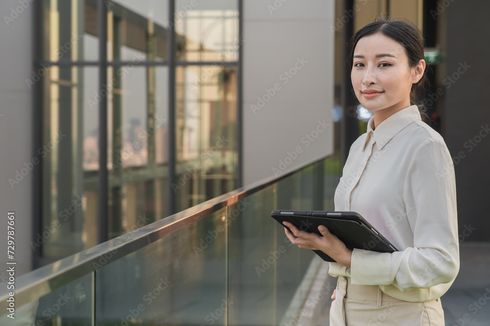 Fototapeta premium Asian woman using digital tablet look in distance planning or visualizing future success, dreaming or thinking, standing on rooftop at sunset. Business vision concept.