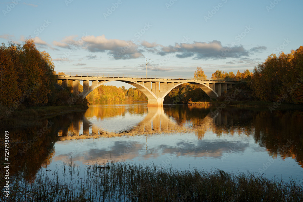 A railway bridge over calm water lit by the sun on a autumn evening with reflection on the water surface
