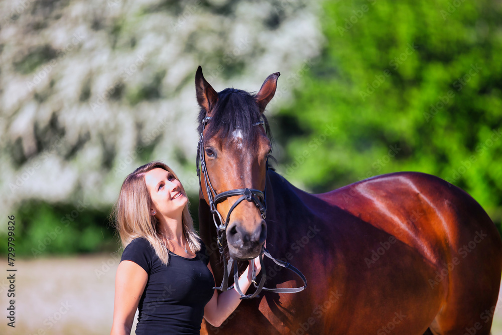 Obraz premium Young brunette woman in the sunshine with her horse in front of white flowering bushes.