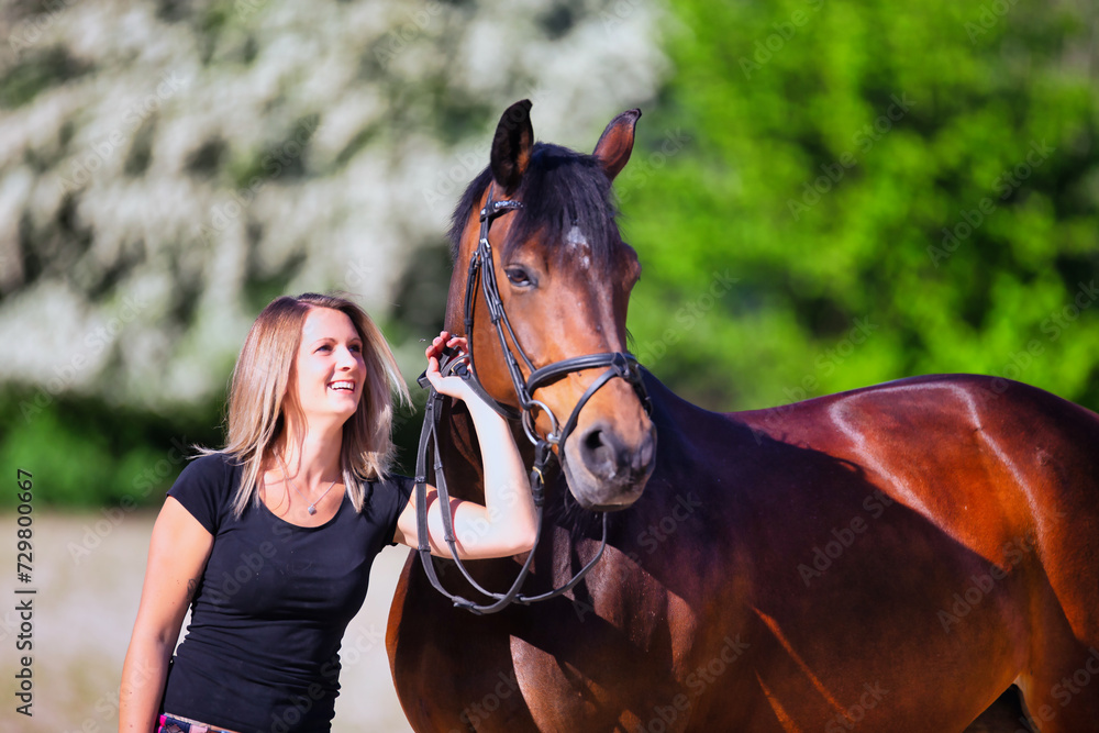 Obraz premium Young brunette woman in the sunshine with her horse in front of white flowering bushes.