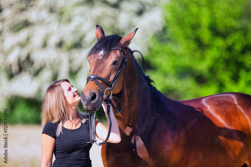 Obraz premium Young brunette woman in the sunshine with her horse in front of white flowering bushes.