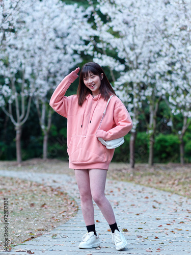 Beautiful young Chinese woman smiling under blooming cherry tree, wearing pink loose oversized top with small white shoulder bag. Candid moment. Emotions, people, beauty, youth and lifestyle portrait.