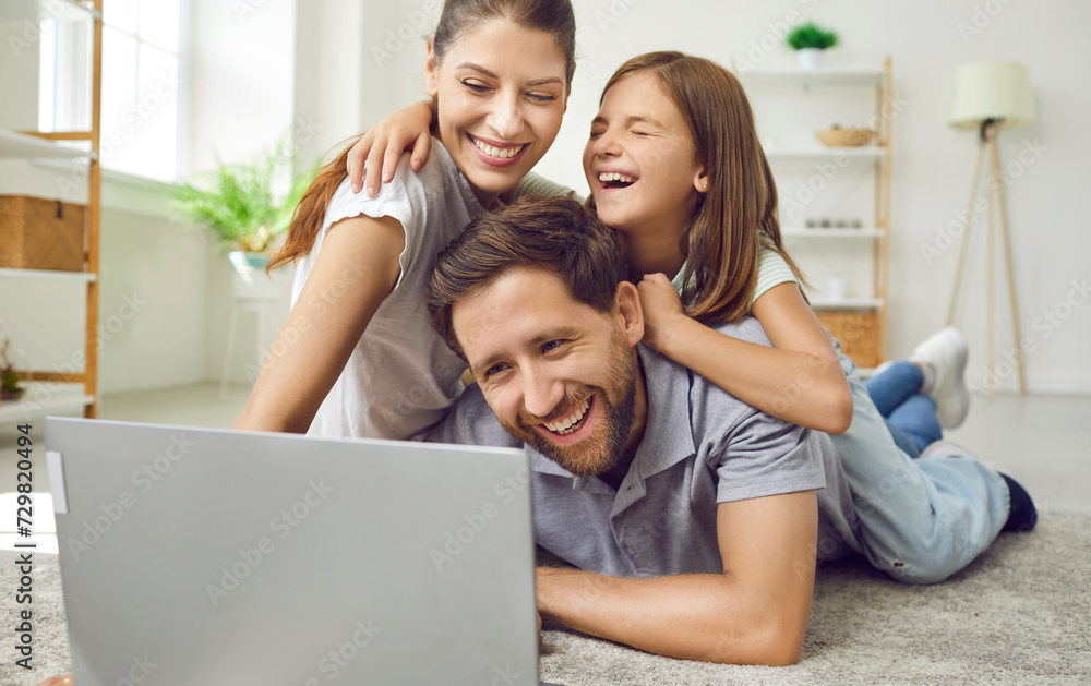 Portrait of happy loving family using laptop while spending time together at home in living room. Mom, dad and their daughter are lying on carpet and watching video, movie or browsing Internet. Portrait of happy loving family using laptop while spending time together at home in living room. Mom, dad and their daughter are lying on carpet and watching video, movie or browsing Internet.