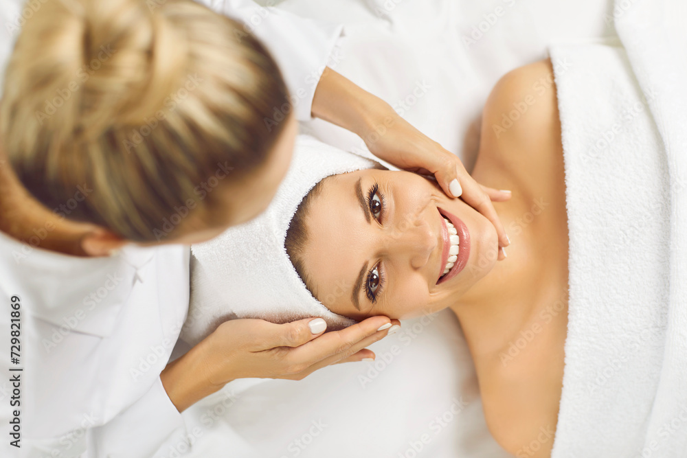 Top view portrait of happy smiling young pretty woman client looking cheerful at camera while lying in spa salon having a face massage and receiving beauty procedure from beautician in beauty clinic.