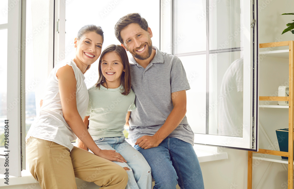 Happy together. Portrait of young married couple and their preteen daughter posing together by window in living room. Nice Caucasian family of three is smiling happily while looking at camera. Happy together. Portrait of young married couple and their preteen daughter posing together by window in living room. Nice Caucasian family of three is smiling happily while looking at camera.