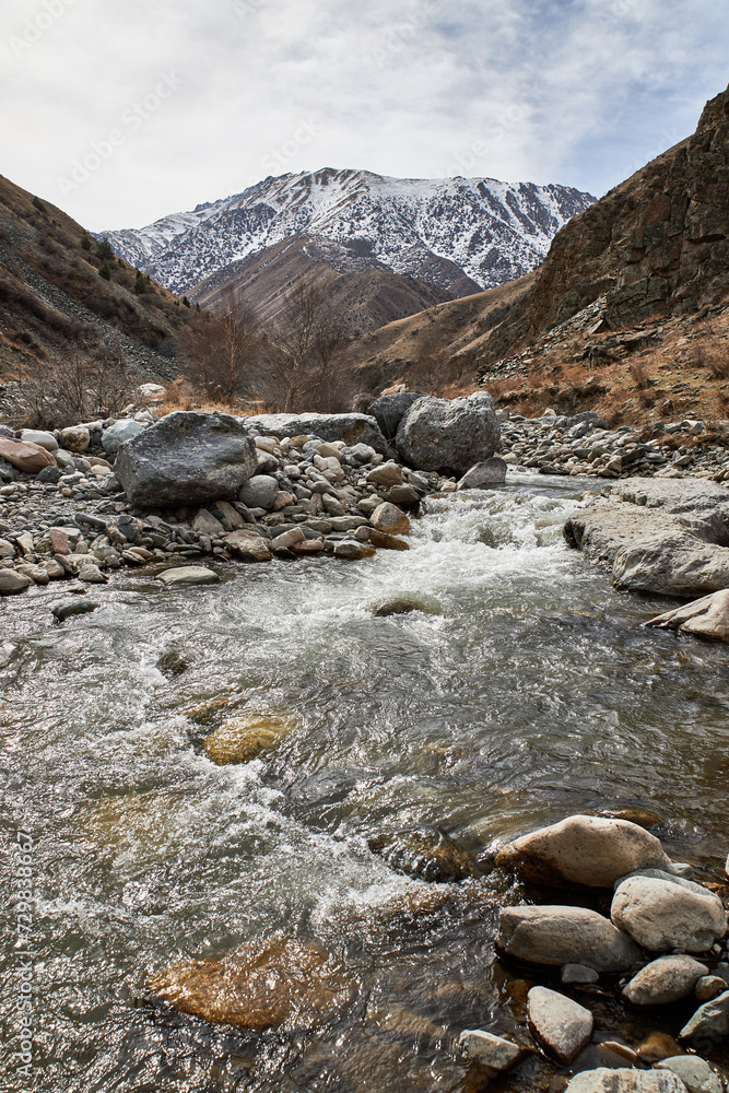 Early spring natural landscape. Snow capped mount, hillsides, large boulders and rocky bank of a shallow river with fast flowing water. Kyrgyz Republic Kyrgyzstan mountains Early spring natural landscape. Snow capped mount, hillsides, large boulders and rocky bank of a shallow river with fast flowing water. Kyrgyz Republic Kyrgyzstan mountains