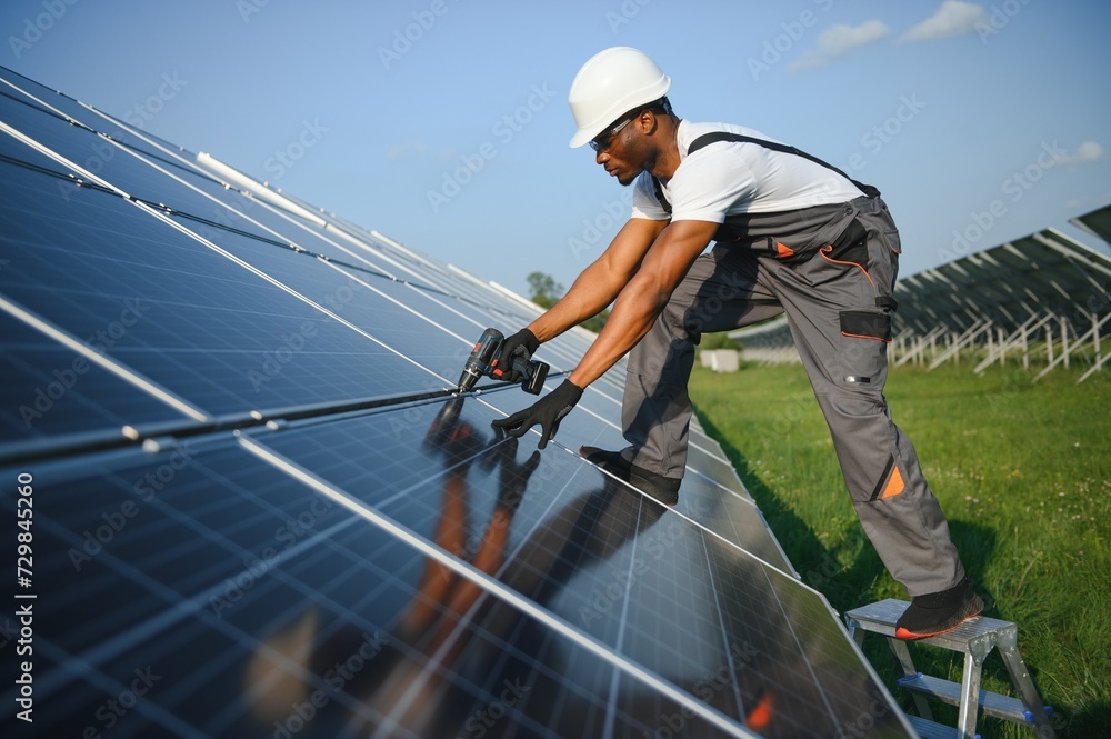 African american man in safety helmet and glasses tighten nuts on solar panels with screwdriver. Competent technician using tools while performing service work on station African american man in safety helmet and glasses tighten nuts on solar panels with screwdriver. Competent technician using tools while performing service work on station