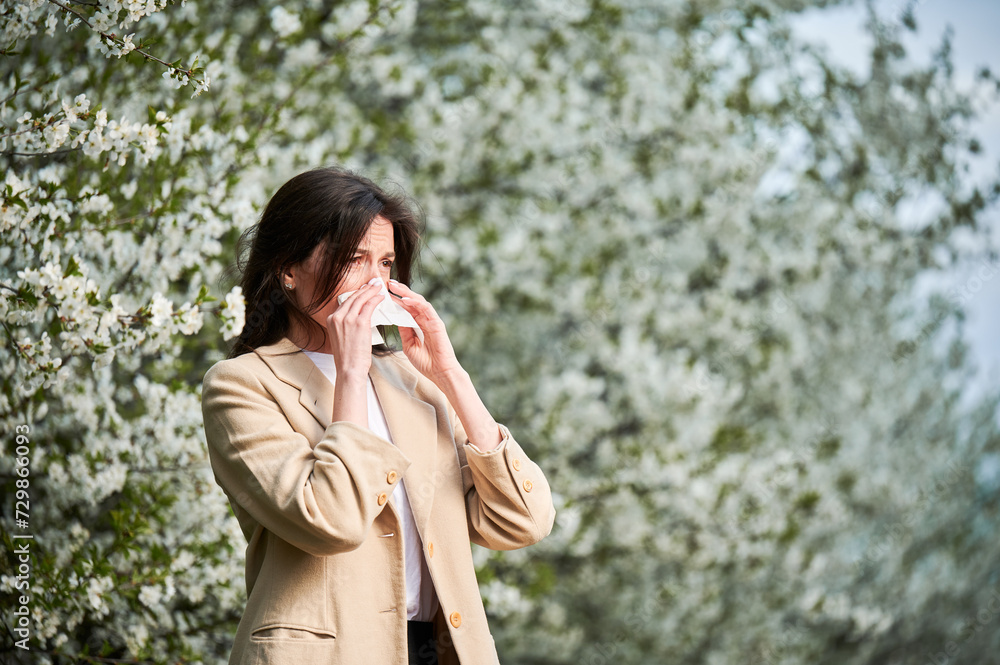 Woman allergic suffering from seasonal allergy at spring in blossoming garden at springtime. Young woman sneezing and blowing nose with nasal handkerchief in front of blooming tree. Allergy concept. Woman allergic suffering from seasonal allergy at spring in blossoming garden at springtime. Young woman sneezing and blowing nose with nasal handkerchief in front of blooming tree. Allergy concept.