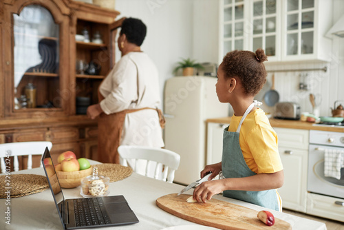 Selective focus shot of African American grandma and granddaughter spending time together cooking meal in kitchen photo