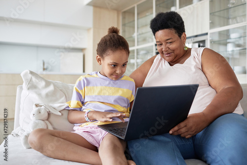 African American grandma and her gen Alpha grandchild sitting on couch in living room surfing Internet on laptop photo