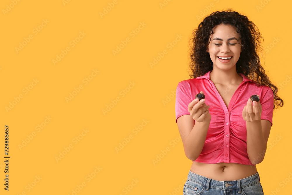 Happy young African-American woman with sweet chocolate candies on yellow background Happy young African-American woman with sweet chocolate candies on yellow background