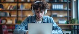 Student watching online lecture using laptop and headphones, taking notes in notebook. Curly-haired guy in office wearing earphones. Close-up. High-quality picture.