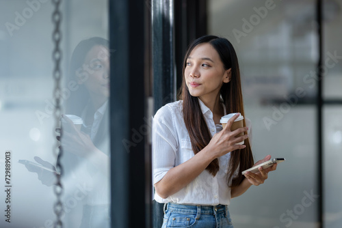A cheerful young businesswoman engaging in a phone conversation while holding a coffee cup, inside a bright modern office space..