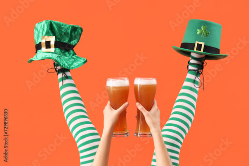 Female legs in green stockings with glasses of beer and leprechaun's hats on orange background. St. Patrick's Day celebration