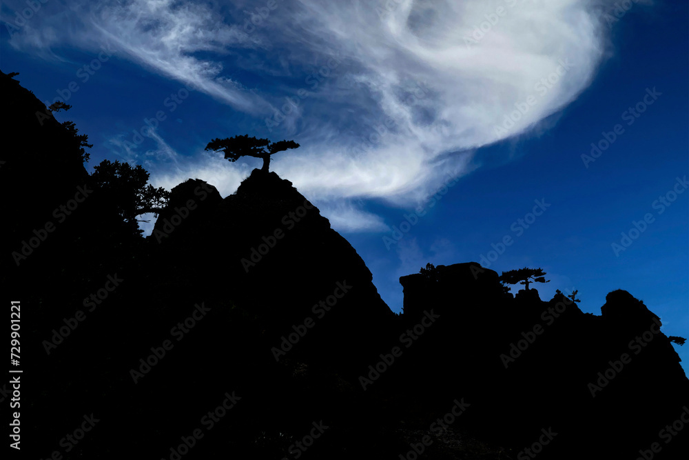 The contour of the shadow of a mountain range with a peak and trees on top against a blue sky with clouds. Mountain landscape. Double exposure.