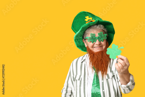 Senior woman in leprechaun hat and decorative glasses with red beard holding clover on yellow background. St. Patrick's Day celebration